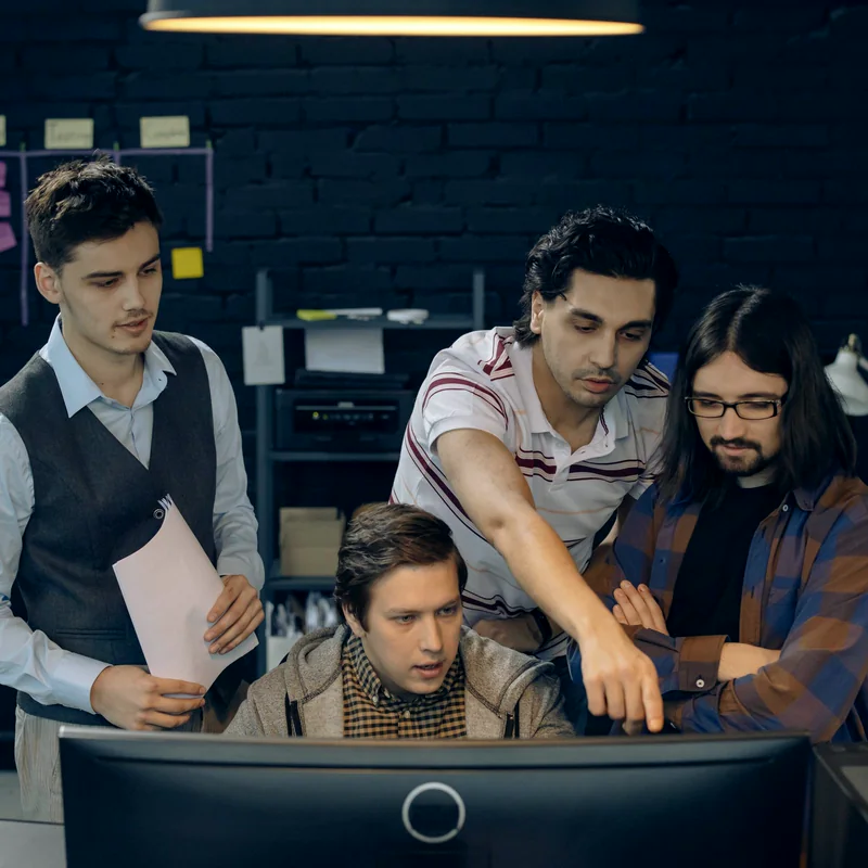 A development team gathered around a monitor, reviewing work together as one colleague points at the screen.