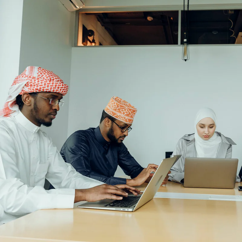Four colleagues in a Gulf-region office working together over laptops and documents around a meeting table.
